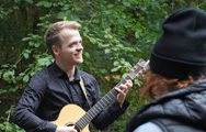 A man plays guitar in a park to a woman and child in a push chair